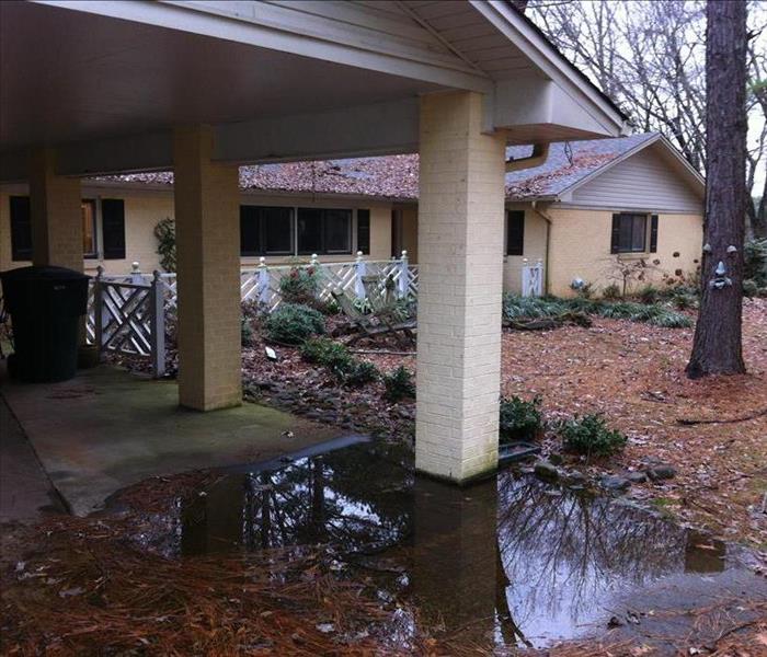 Water covers the front yard of a home. Several fallen tree branches and leaves lay on the grass.