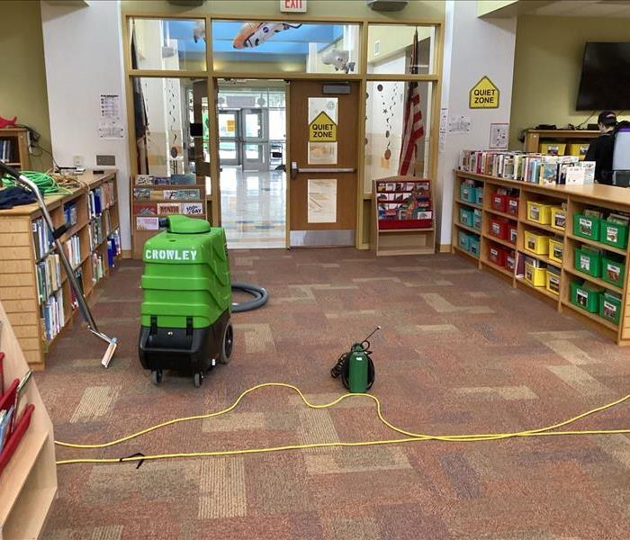 Green carpet cleaner and sprayer in elementary school library with technician vacuuming in background